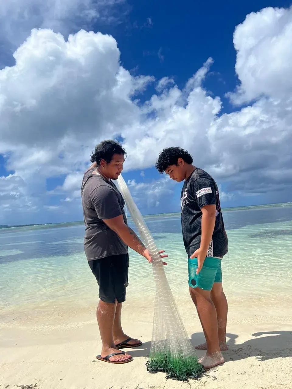 Two people standing in the shallows, one holding a talåya net, under a bright sky