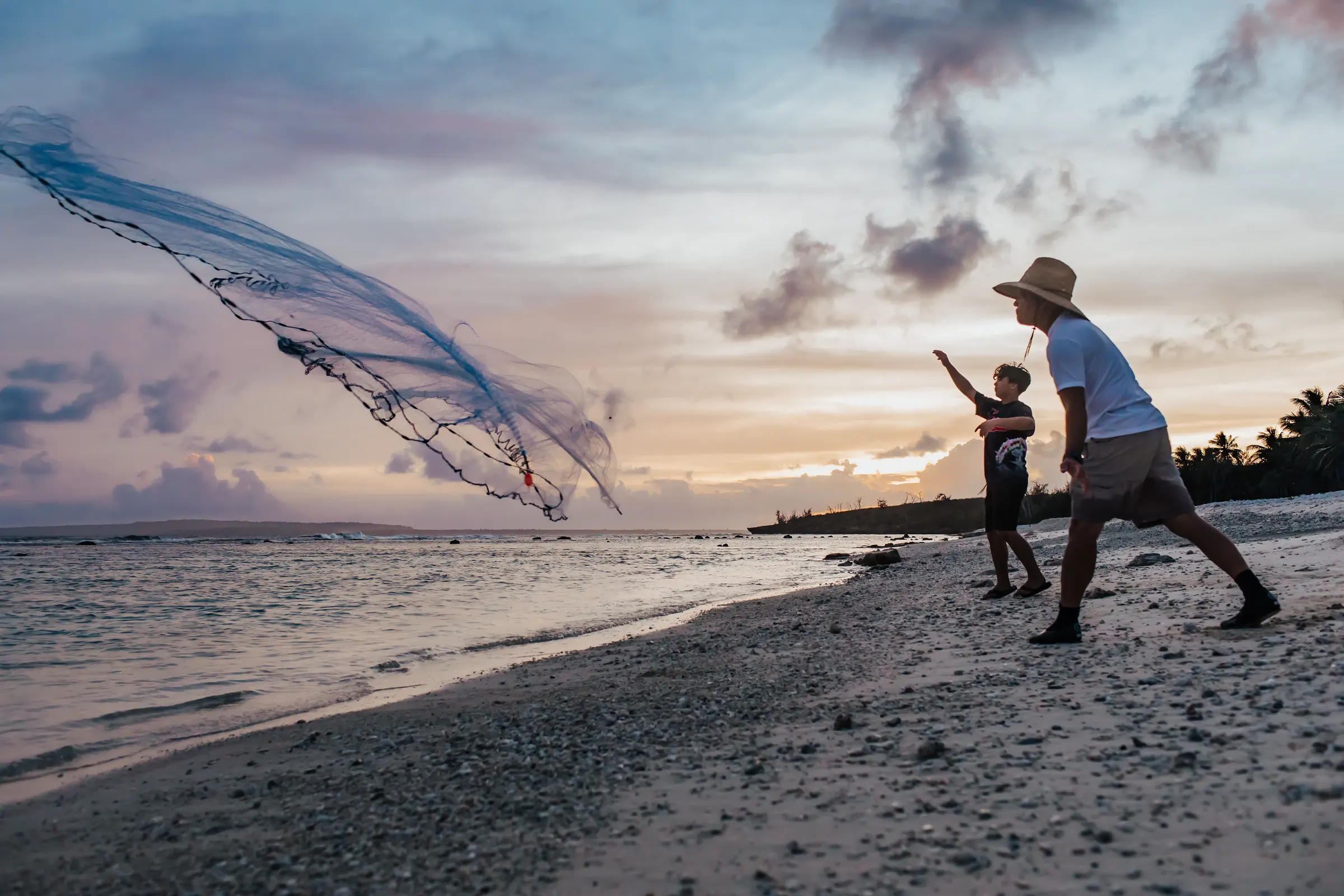 A talåya net opening mid-air over the water at sunset as a fisher casts from the beach